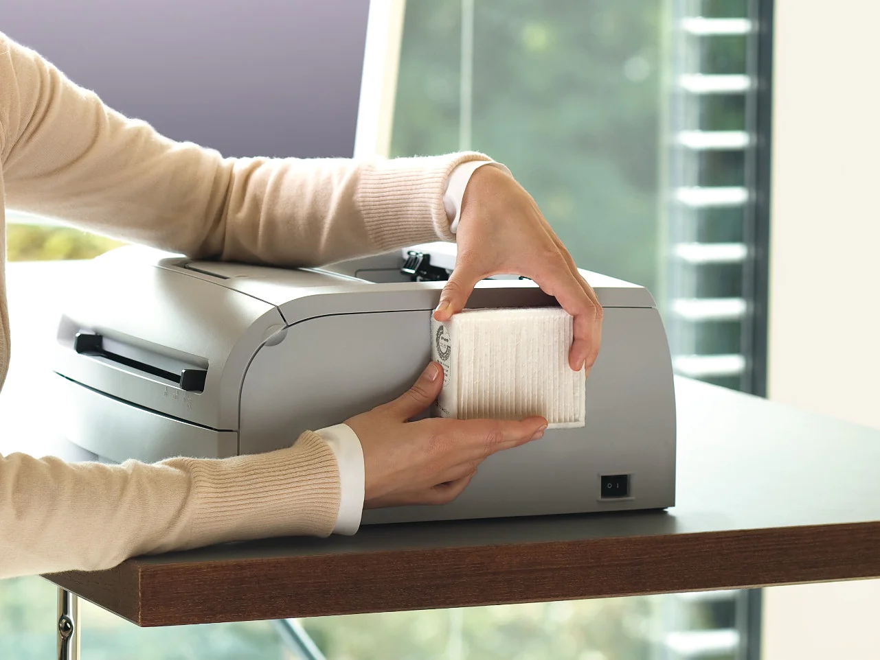 A person with blonde hair is seated at a desk, using a printer. They are smiling and removing a printed sheet from the device, with tesa tape on hand for any quick fixes needed. There is a computer monitor in the background. Sunlight streams through large windows with a view of green trees. (This text has been generated by AI)