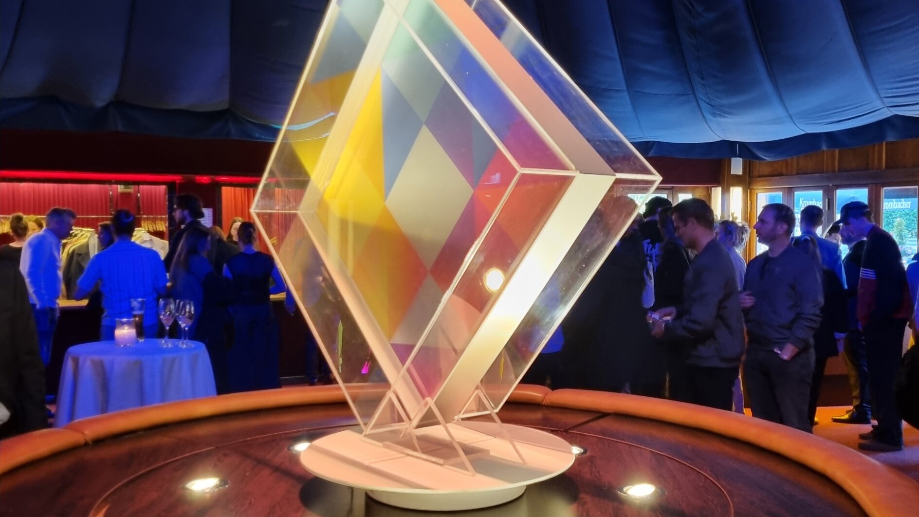 A geometric glass sculpture with colorful panels stands on a round wooden platform under spotlights, while people in formal attire gather and converse in the background of a dimly lit event space. (This text has been generated by AI)