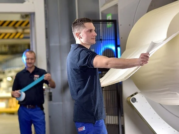 Two workers in a factory handle large rolls of white material. One man in the foreground inspects the sheet, while another in the background holds a roll of blue tesa tape. Both wear dark shirts and blue pants. Industrial equipment is visible. (This text has been generated by AI)