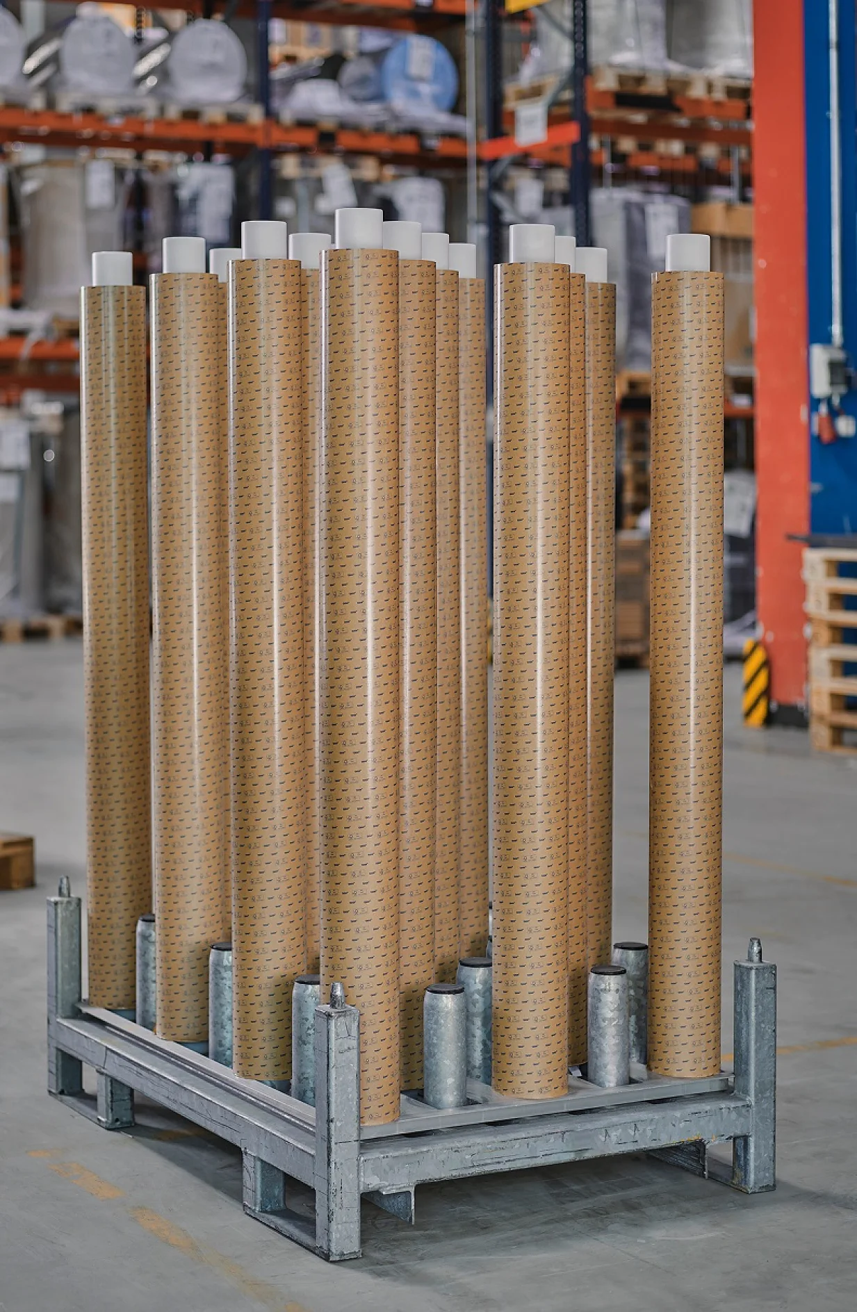A metal pallet holds multiple upright cylindrical rolls of brown industrial tesa tape material in a warehouse; shelves and other equipment are visible in the background. (This text has been generated by AI)