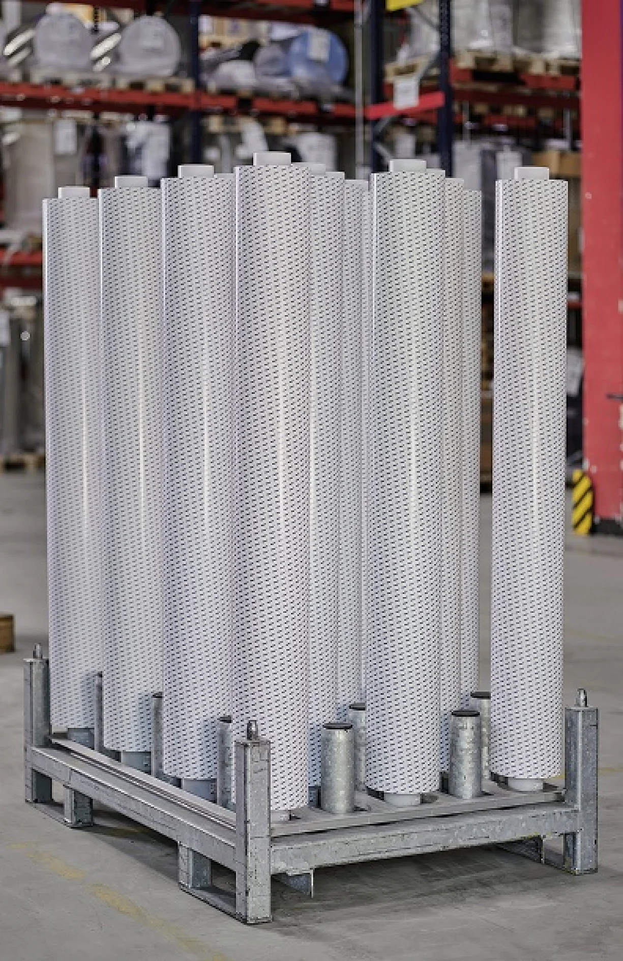 A metal pallet holds ten vertically standing, perforated cylindrical metal tubes in a warehouse with shelves and industrial equipment in the background. Each tube is secured using tesa tape. (This text has been generated by AI)