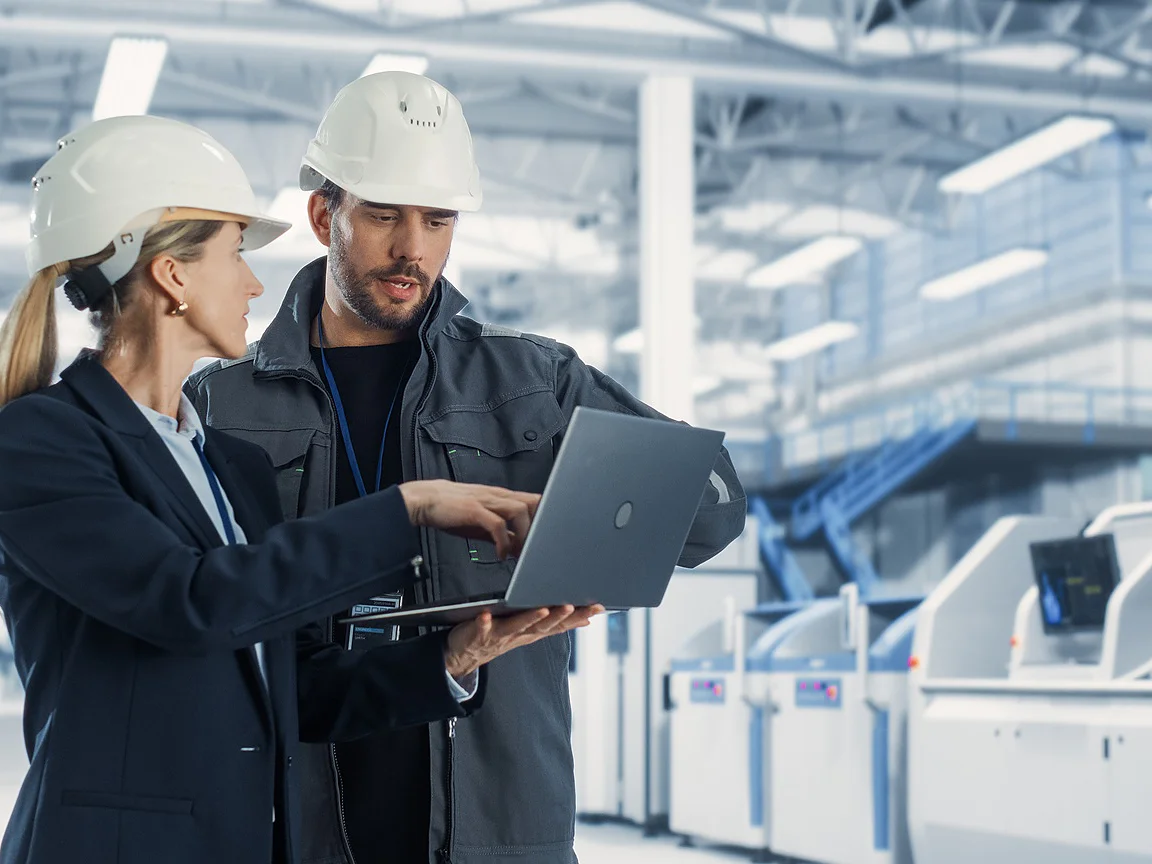 Two people in safety helmets and work attire stand in an industrial facility, looking at and discussing information on a laptop. Industrial machines, bright lighting, and tesa tape are visible in the background. (This text has been generated by AI)