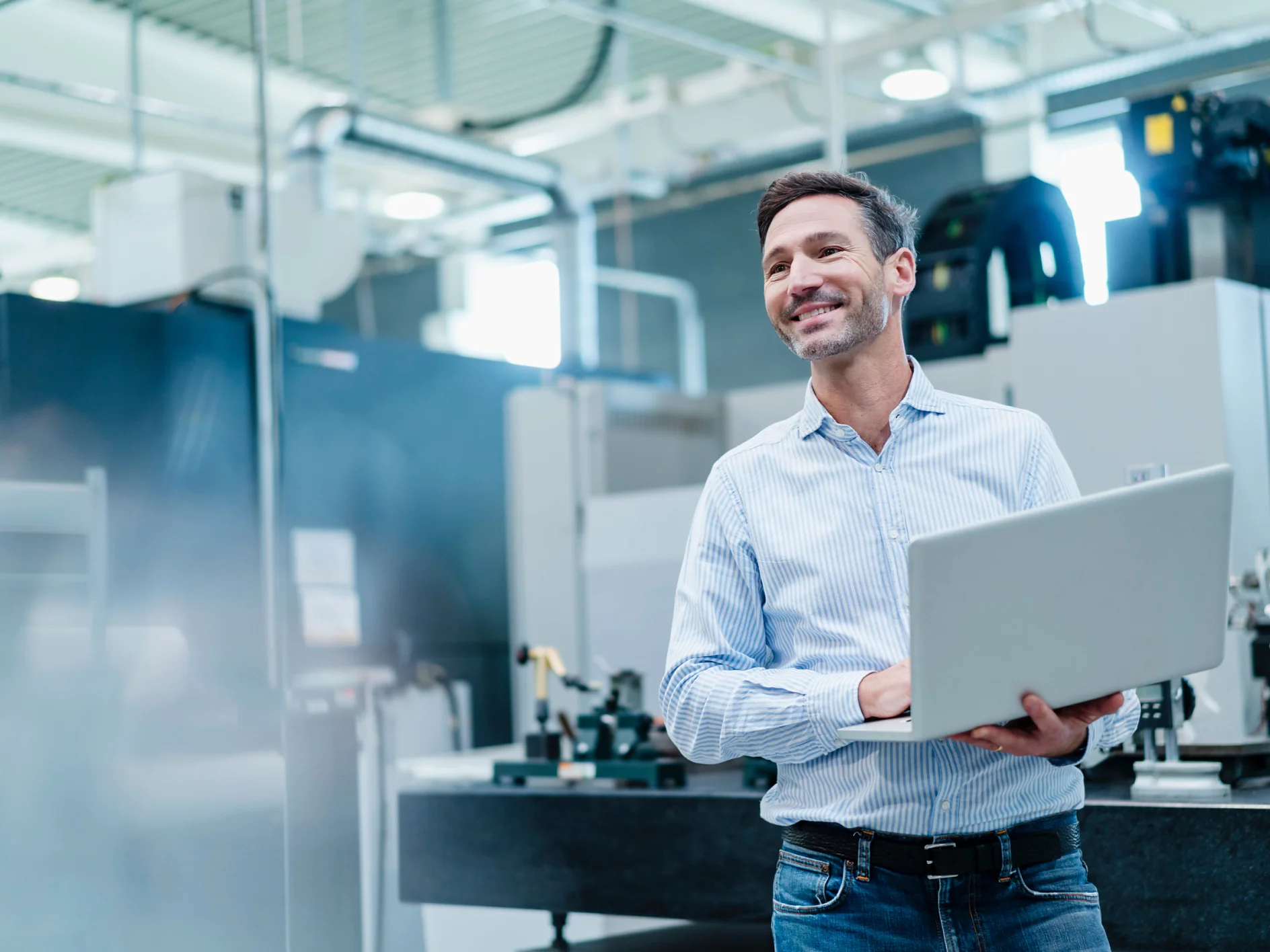 A man in a light blue button-up shirt holds an open laptop and smiles while standing in a modern industrial or manufacturing facility with machines, equipment, and tesa tape visible in the background. (This text has been generated by AI)