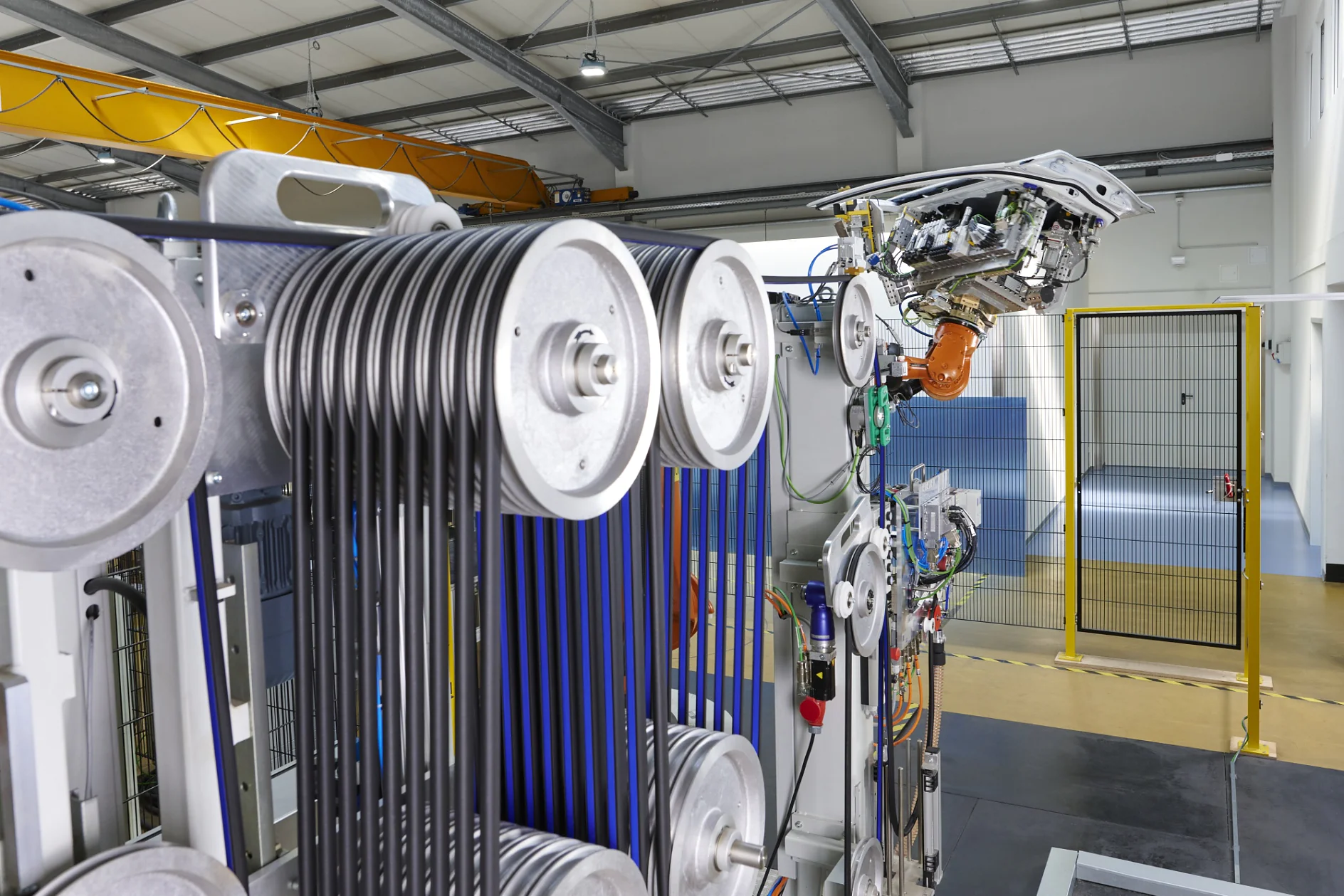 A close-up view of an industrial machine with multiple large metal pulleys and black belts secured using tesa tape inside a factory setting. The background shows metal fencing, a blue wall, and a yellow overhead crane. (This text has been generated by AI)