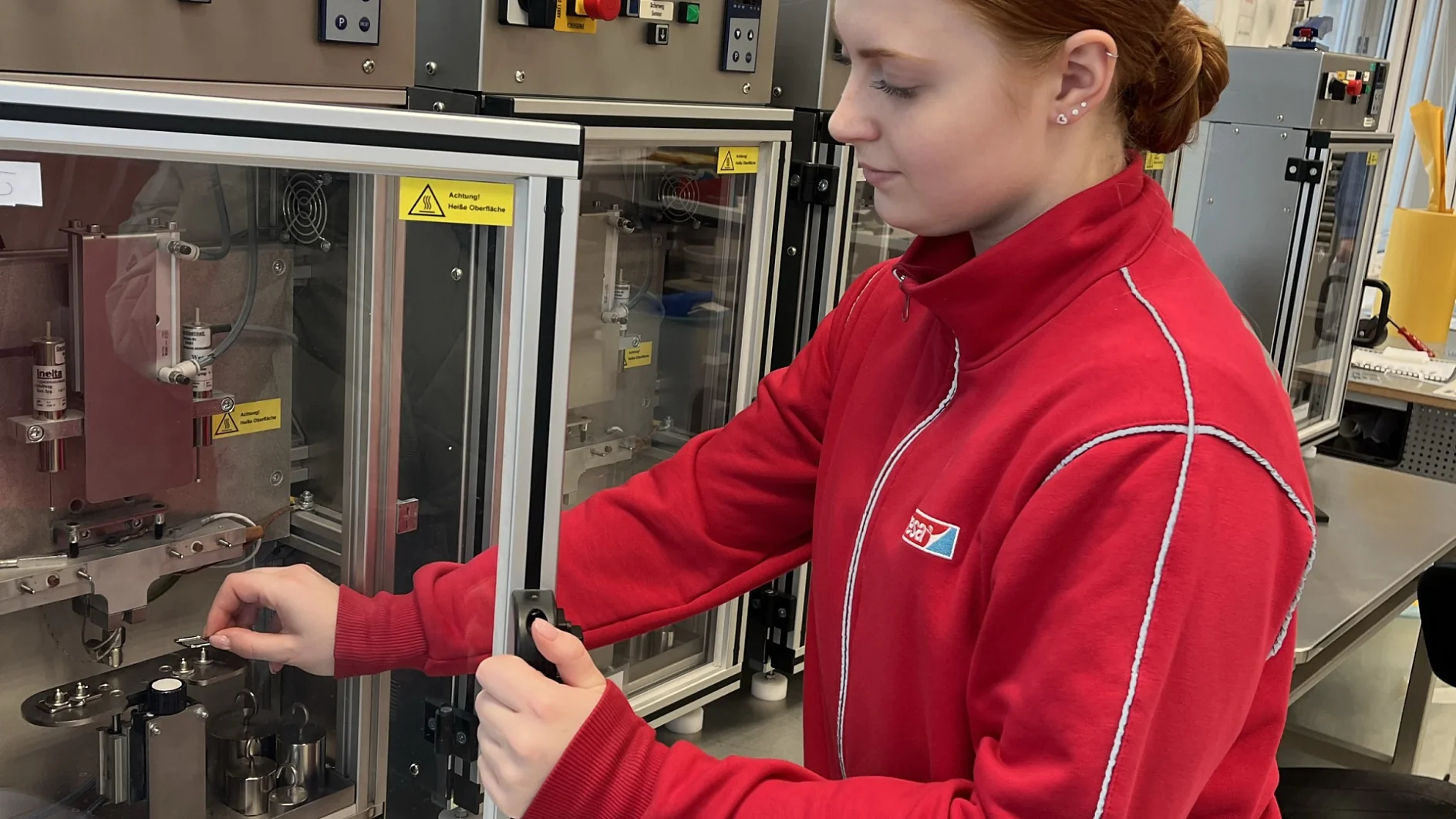 A person in a red jacket operates scientific equipment inside a laboratory. They are adjusting controls on a machine with glass doors and various switches, standing at a metal counter with windows in the background. (This text has been generated by AI)