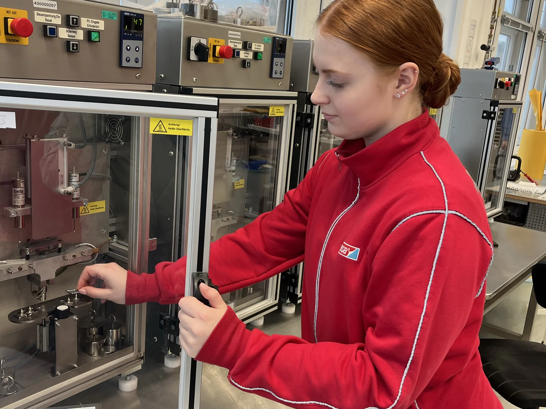 A person in a red jacket operates scientific equipment inside a laboratory. They are adjusting controls on a machine with glass doors and various switches, standing at a metal counter with windows in the background. (This text has been generated by AI)