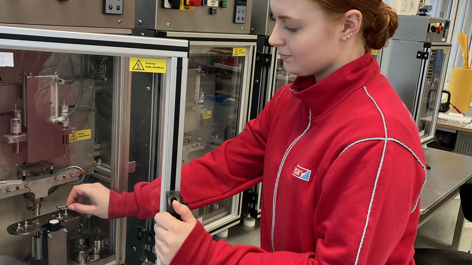 A person in a red jacket operates scientific equipment inside a laboratory. They are adjusting controls on a machine with glass doors and various switches, standing at a metal counter with windows in the background.