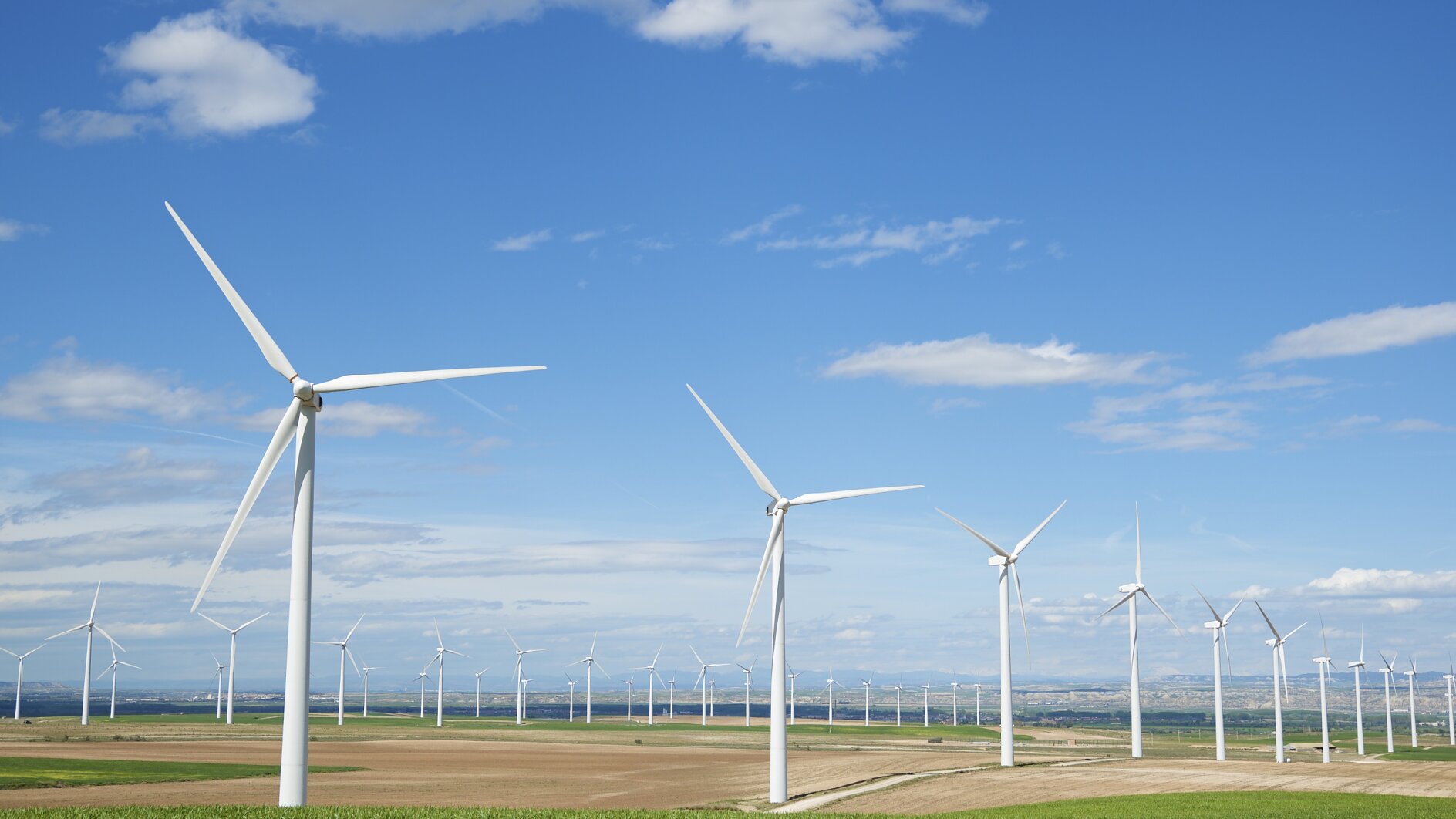 Several white wind turbines stand in rows across a flat, open field with green and brown patches under a blue sky with scattered white clouds. (This text has been generated by AI)