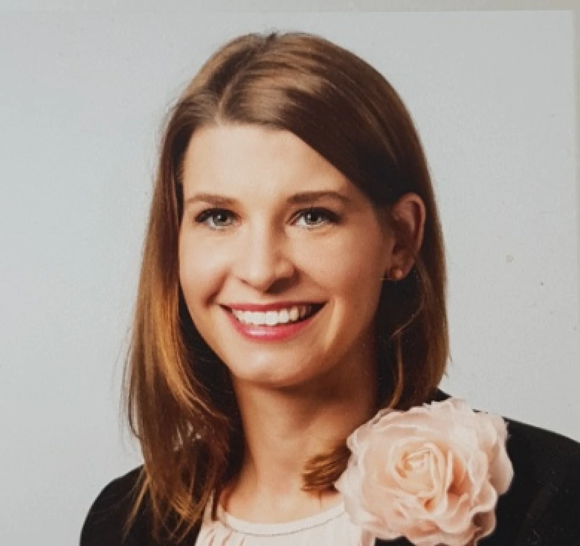A woman with straight, shoulder-length brown hair is smiling. She is wearing a black blazer, a light-colored top, and a large pale pink flower pin on her lapel crafted from tesa tape. The background is plain and light-colored. (This text has been generated by AI)
