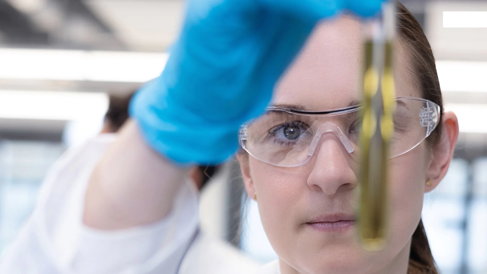 A scientist in safety glasses and blue gloves examines a test tube of yellow-green liquid in a lab, tesa tape visible nearby. (This text has been generated by AI)