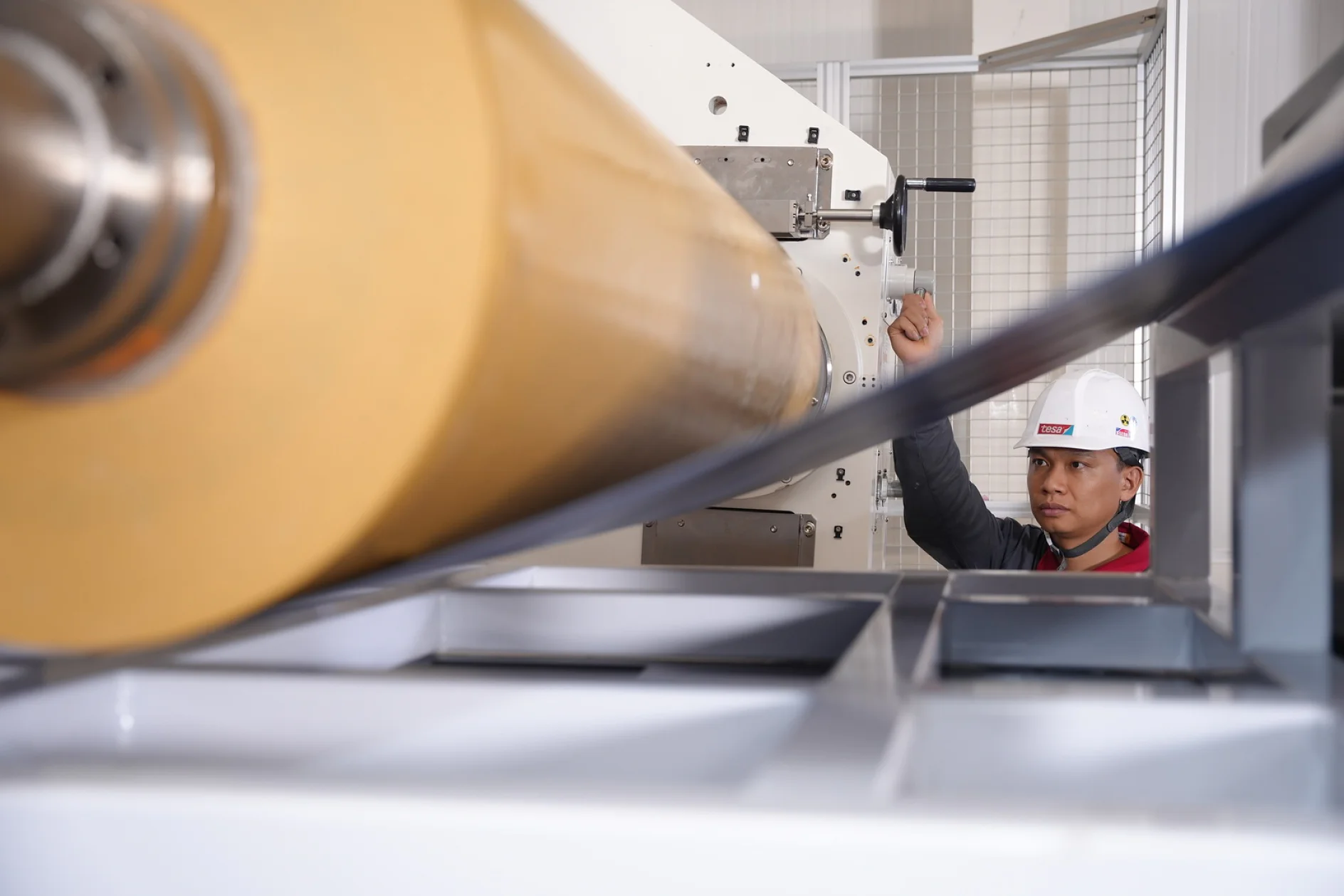 A person in a white safety helmet and gray uniform operates machinery with large yellow and metallic rollers for tesa tape in an industrial setting. (This text has been generated by AI)