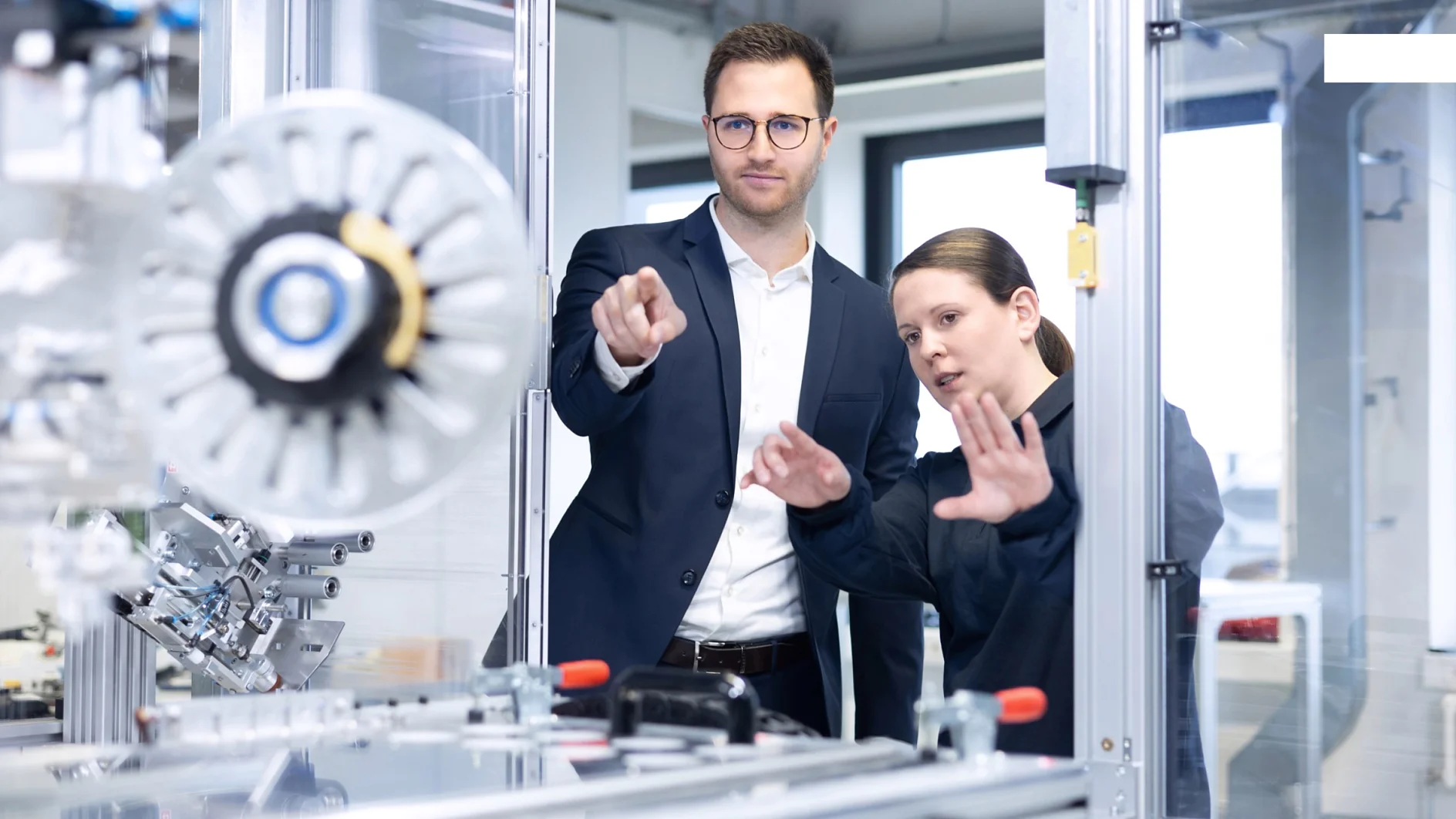 A man in a suit and a woman in work attire stand by industrial machinery in a modern lab, both focused on tesa tape equipment. (This text has been generated by AI)