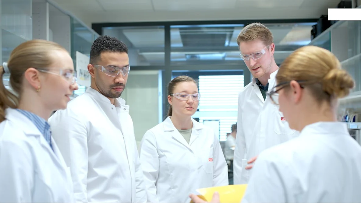 Five people in lab coats and safety glasses stand in a laboratory, gathered around someone holding yellow tesa tape, discussing together. (This text has been generated by AI)