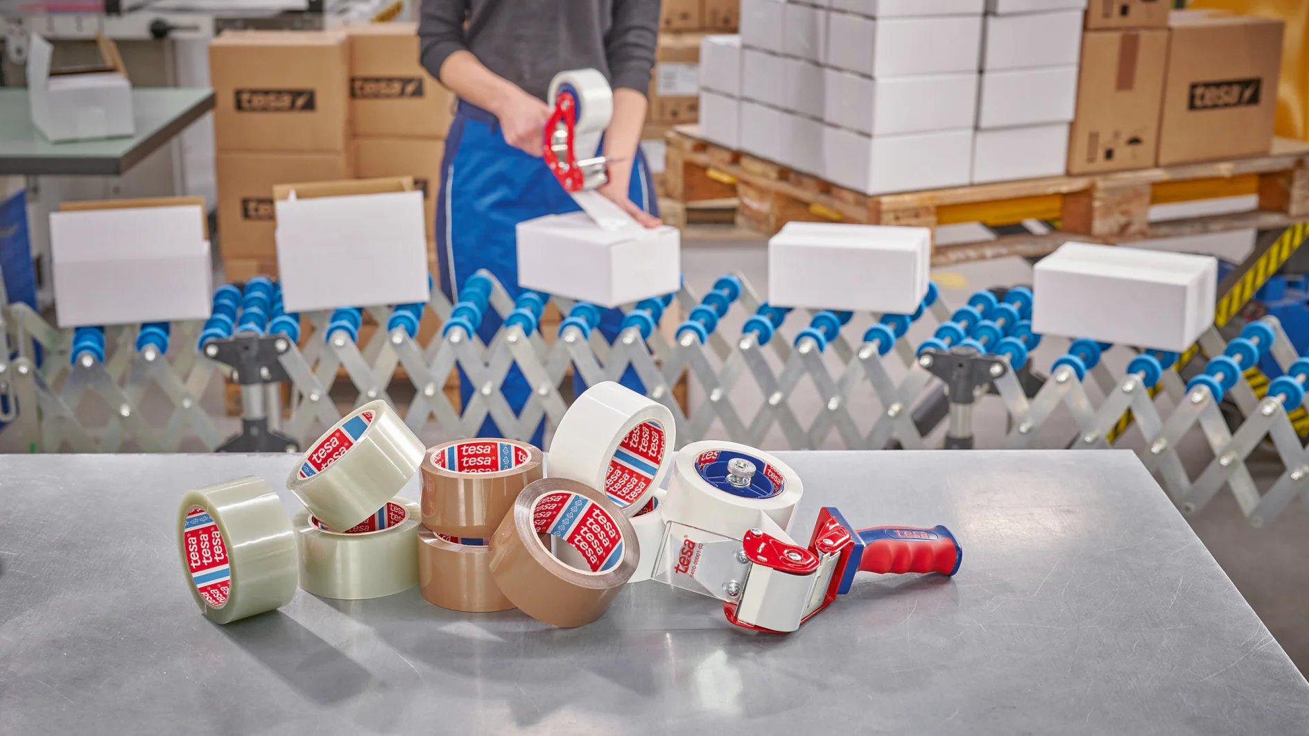 A person seals a cardboard box with a tesa tape dispenser. Rolls of tesa tape, dispensers, boxes, and a conveyor are visible nearby. (This text has been generated by AI)