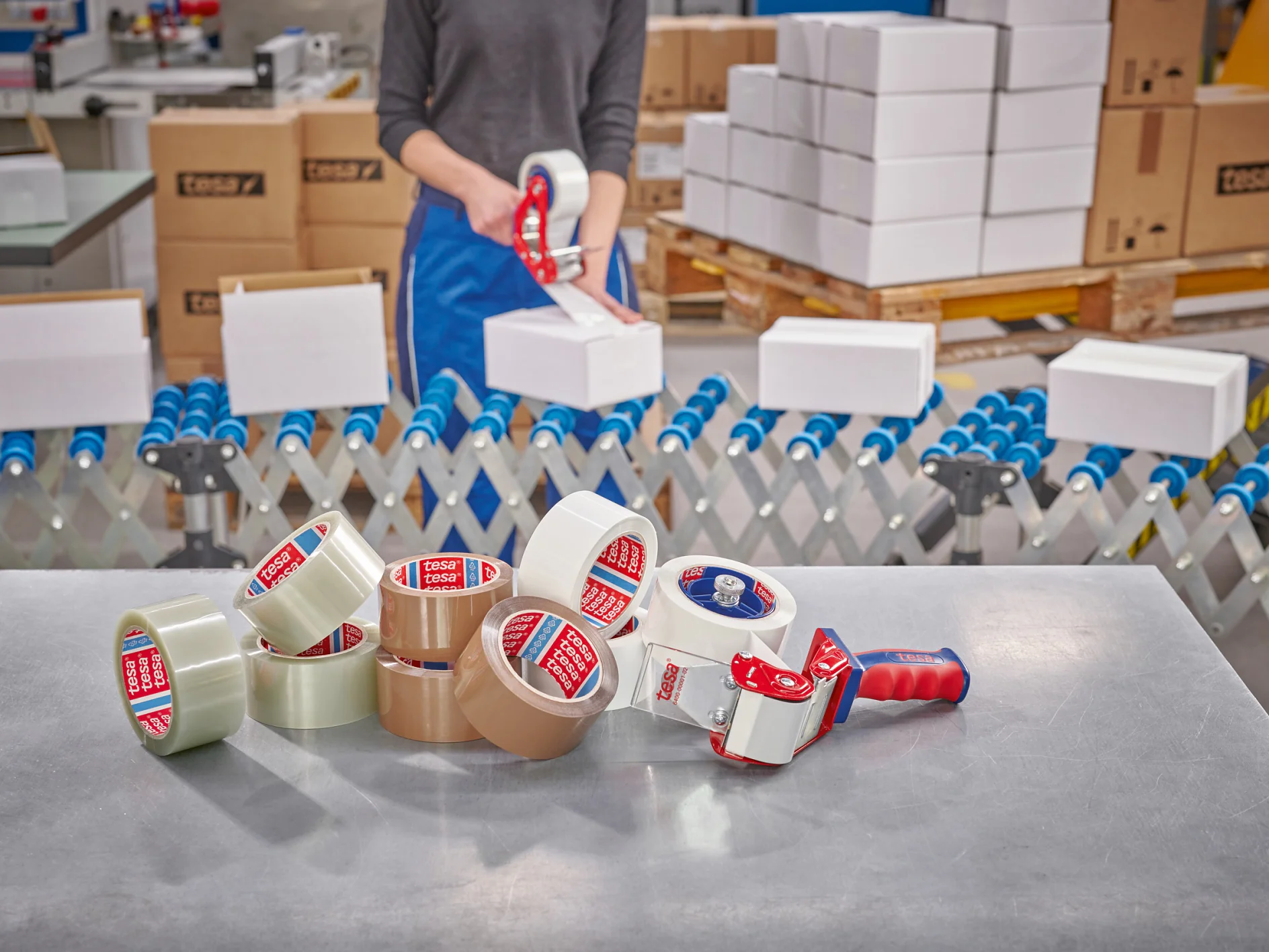 A person seals a cardboard box with a tesa tape dispenser. Rolls of tesa tape, dispensers, boxes, and a conveyor are visible nearby. (This text has been generated by AI)