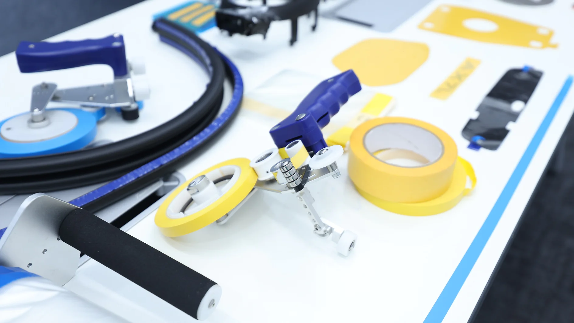 A white table with various tools and materials, including rolls of yellow tesa tape, handles, a blue and silver cutter, black tubing, and small metal parts neatly arranged. (This text has been generated by AI)