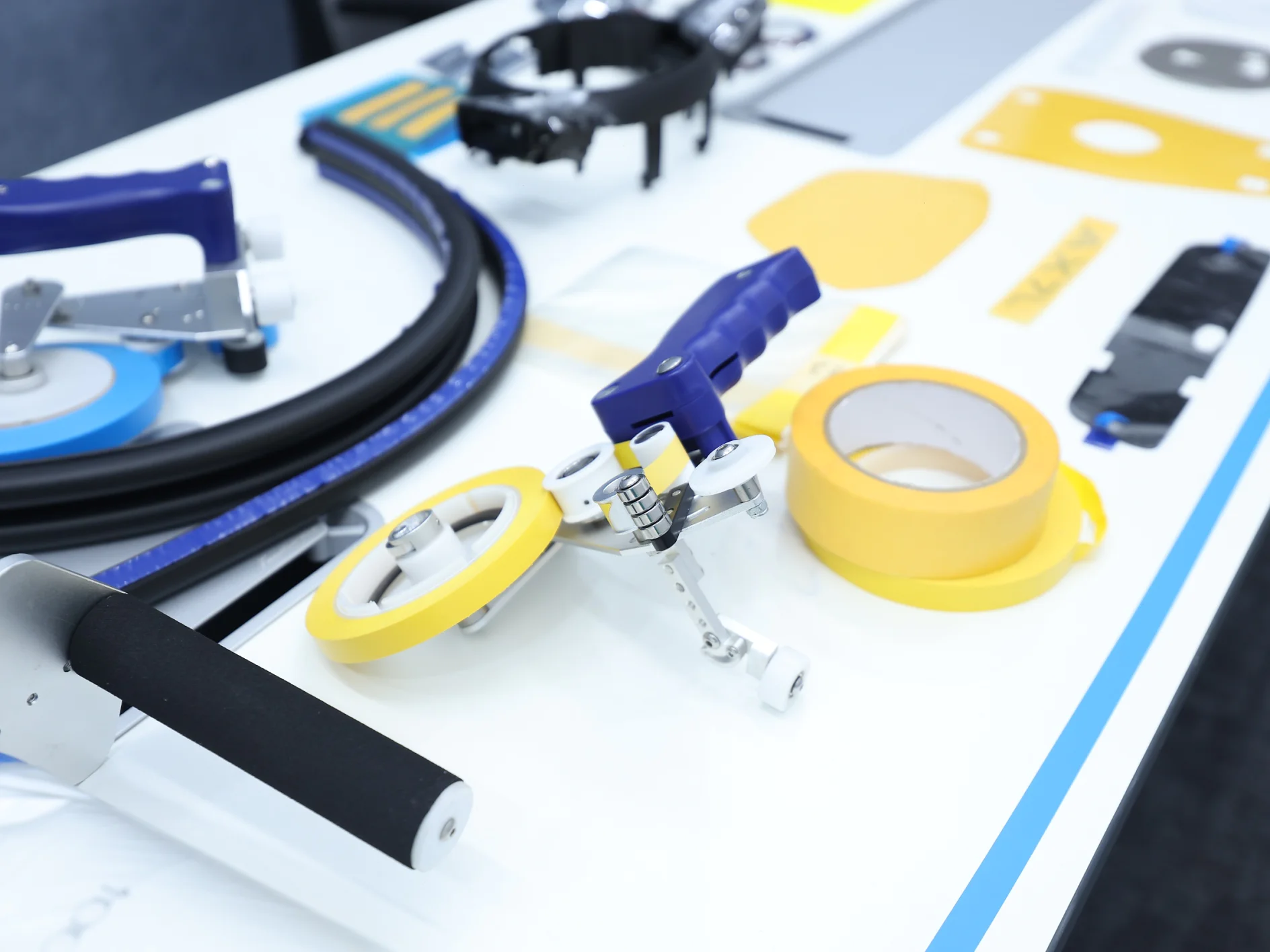 A white table with various tools and materials, including rolls of yellow tesa tape, handles, a blue and silver cutter, black tubing, and small metal parts neatly arranged. (This text has been generated by AI)