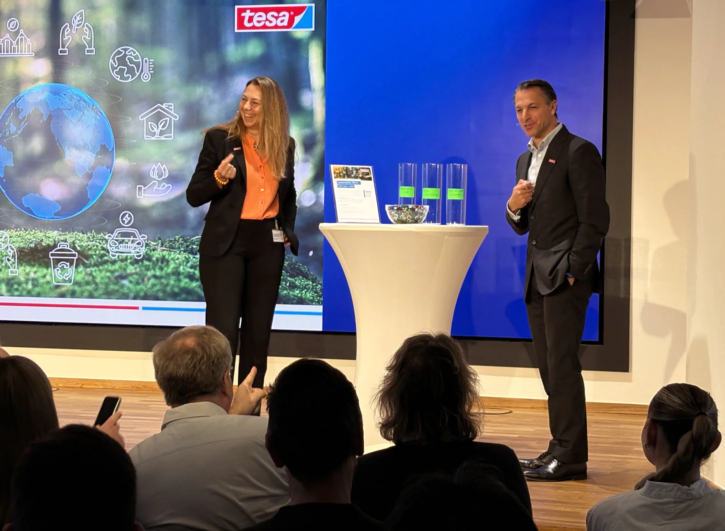 Sustainability Week 2025_Ingrid Sebald + Kourosh Bahrami A woman and a man stand on stage by a table with tesa tape cylinders and a bowl. A tesa logo screen is behind them; audience watches. (This text has been generated by AI)