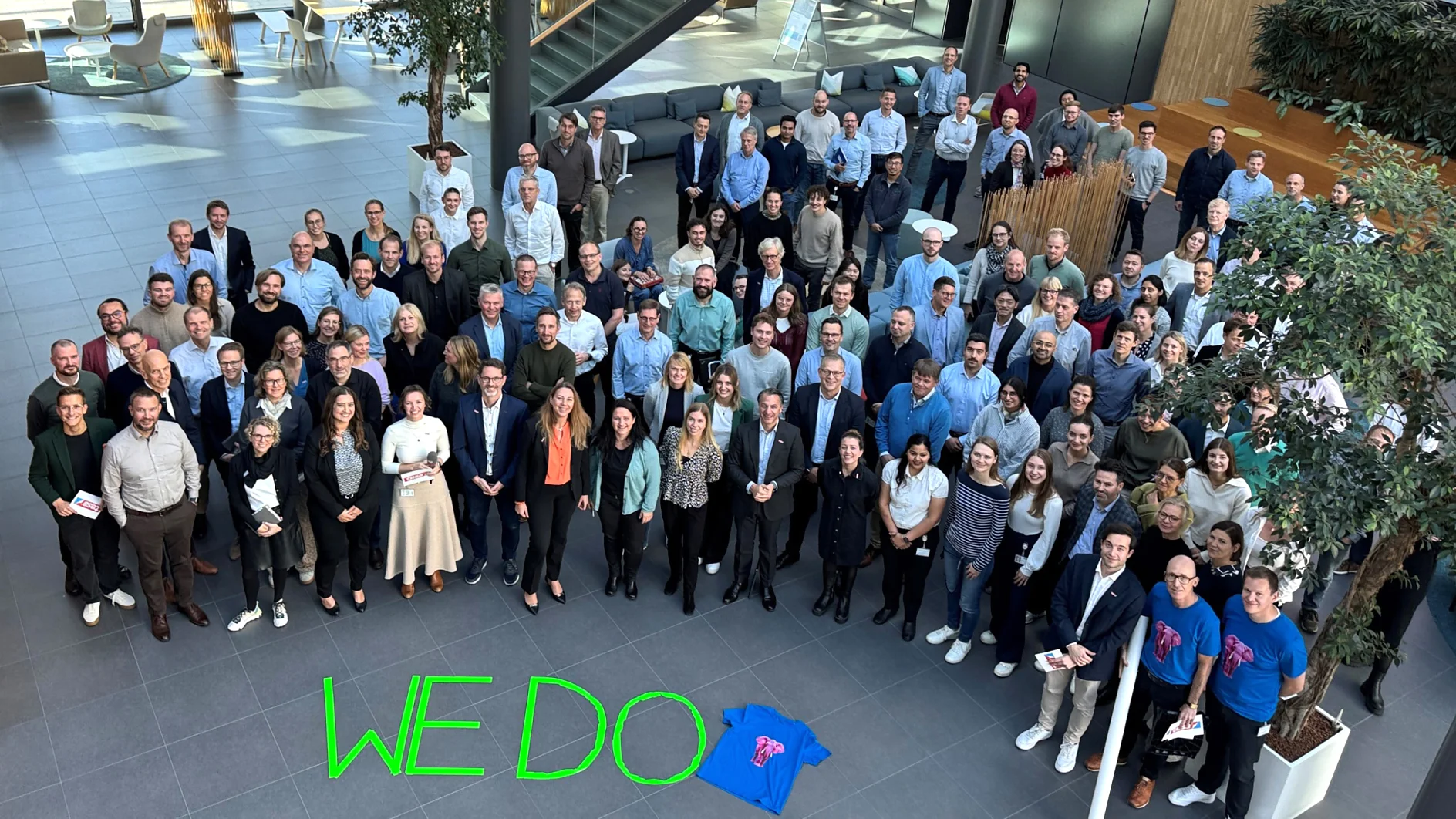 A large group poses in a sunlit atrium. A blue t-shirt and the words "WE DO" in green, written with tesa tape, are in front. (This text has been generated by AI)