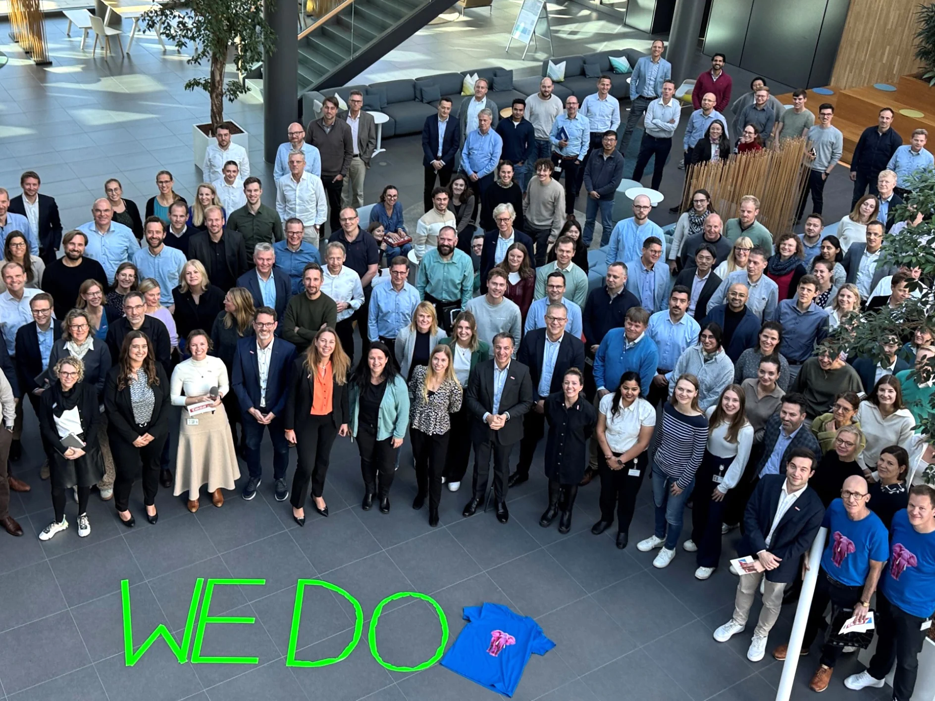 A large group poses in a sunlit atrium. A blue t-shirt and the words "WE DO" in green, written with tesa tape, are in front. (This text has been generated by AI)