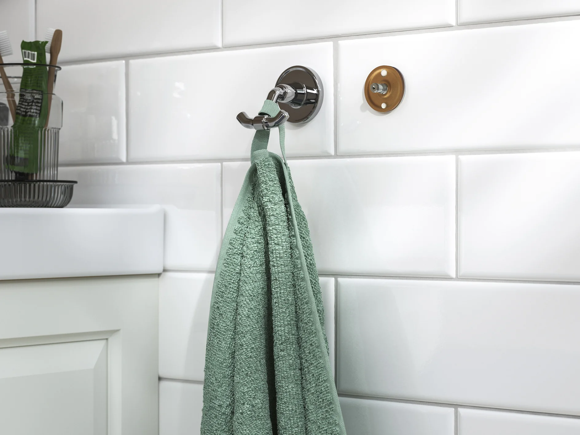 A green hand towel hangs from a chrome wall hook fixed with tesa tape on white subway tiles. Toiletries sit on the left sink countertop. (This text has been generated by AI)