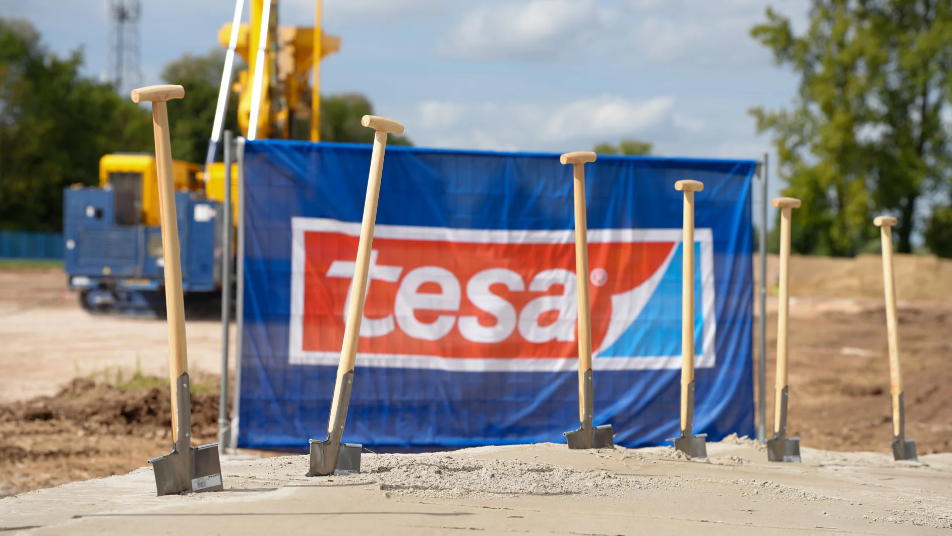 Six shovels stand in a concrete slab at a construction site. A blue tesa tape banner, equipment, trees, and clouds are visible. (This text has been generated by AI)