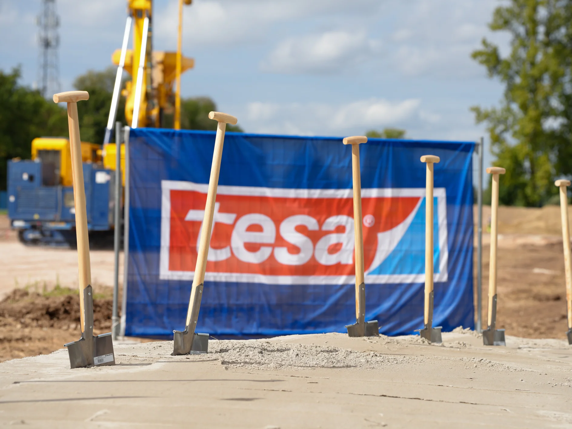 Six shovels stand in a concrete slab at a construction site. A blue tesa tape banner, equipment, trees, and clouds are visible. (This text has been generated by AI)