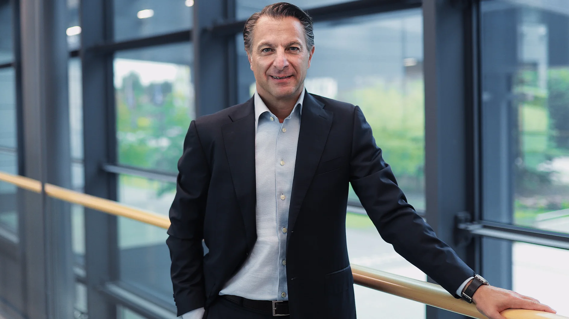 A man in a dark suit and light blue shirt stands by a railing indoors. Large windows show greenery outside. He smiles at the camera. (This text has been generated by AI)