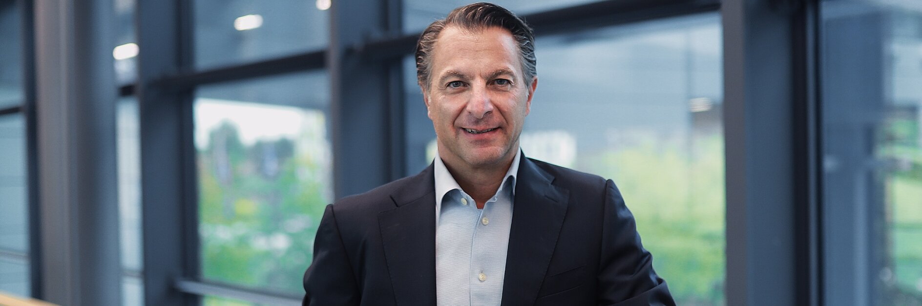 A man in a dark suit and light blue shirt stands by a railing indoors. Large windows show greenery outside. He smiles at the camera. (This text has been generated by AI)