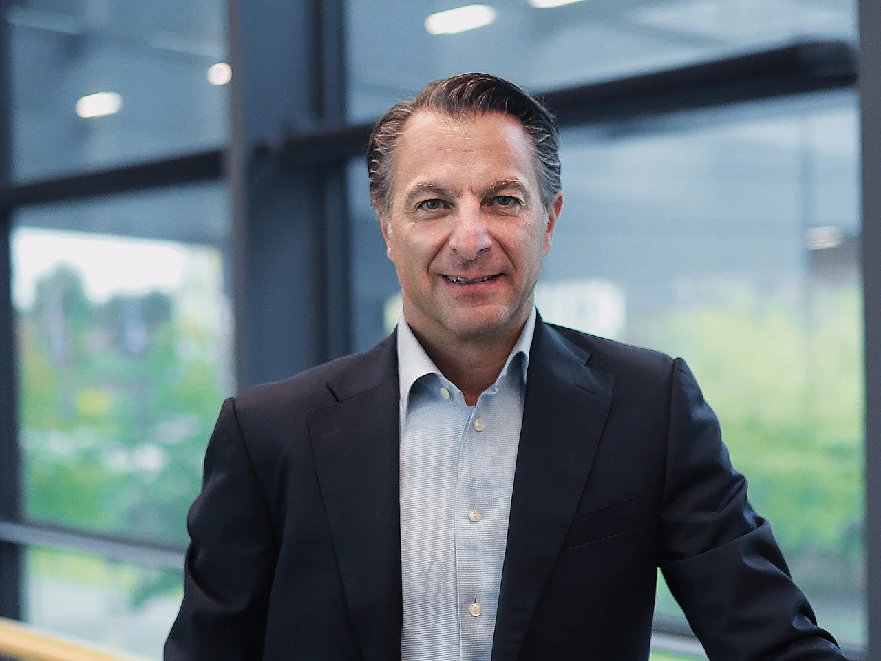 A man in a dark suit and light blue shirt stands by a railing indoors. Large windows show greenery outside. He smiles at the camera. (This text has been generated by AI)
