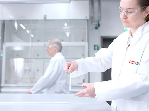 A person in a white lab coat and safety glasses handles a transparent tesa tape object in a bright lab, another in the background. (This text has been generated by AI)