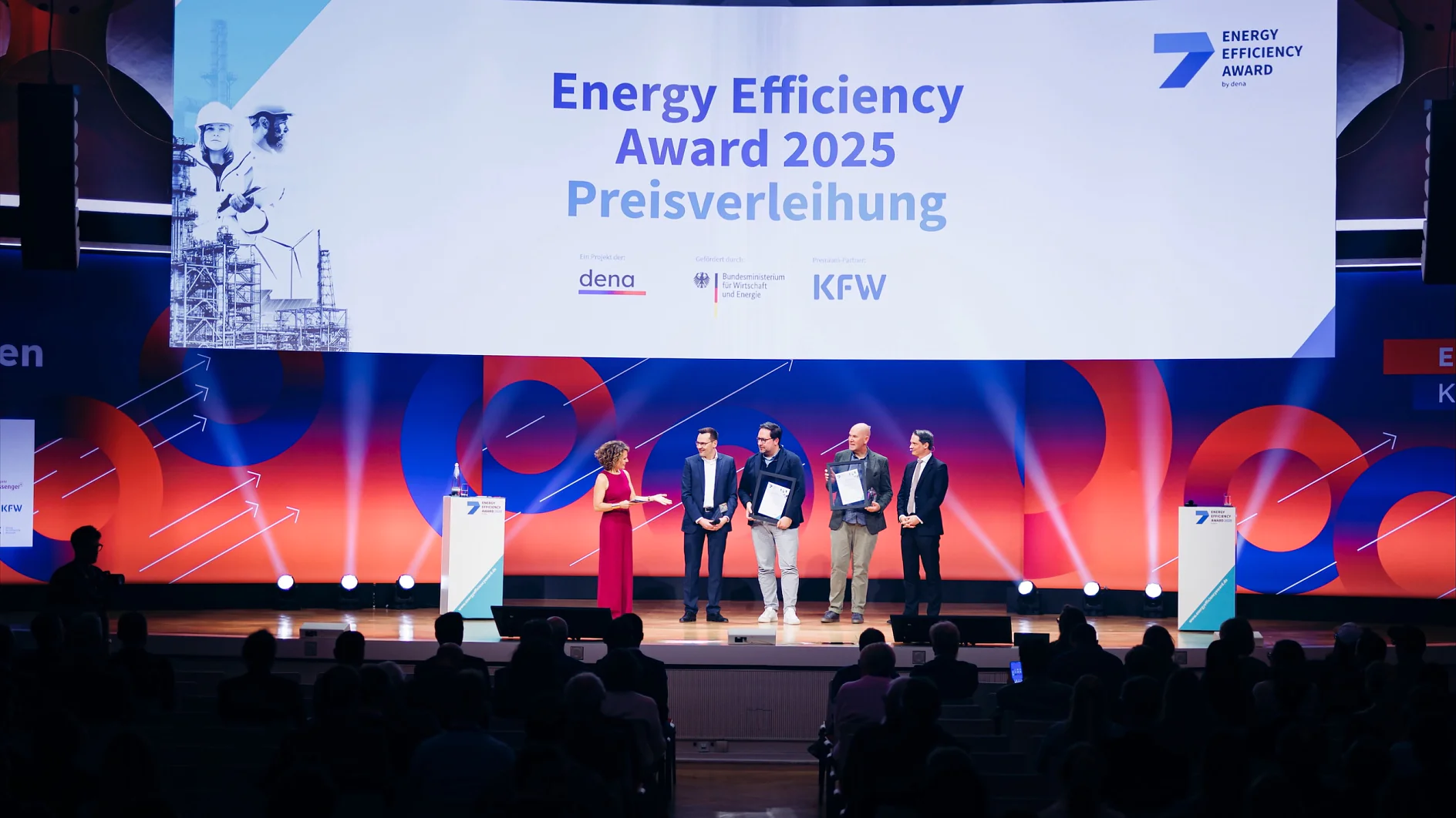 Five people on stage at the Energy Efficiency Award 2025; one speaks, four hold certificates. tesa tape banner behind them. (This text has been generated by AI)