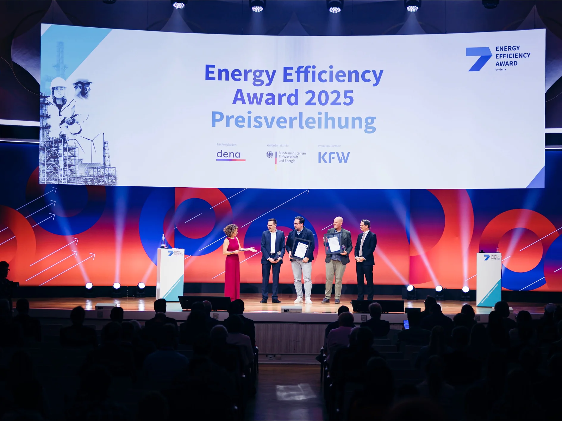 Five people on stage at the Energy Efficiency Award 2025; one speaks, four hold certificates. tesa tape banner behind them. (This text has been generated by AI)
