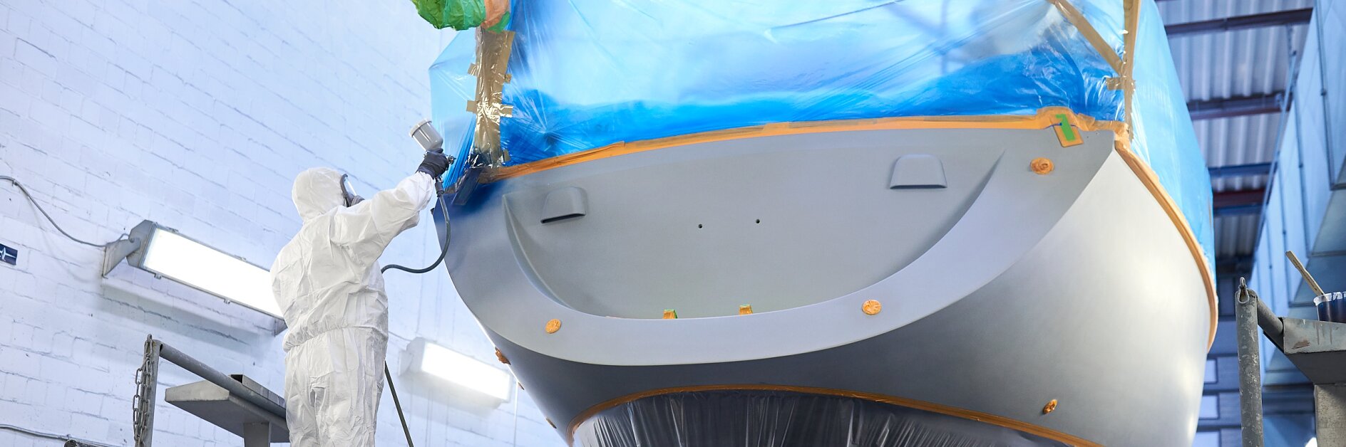 worker in protective suit applying blue tesa tape to boat hull in indoor workshop during maintenance or repair