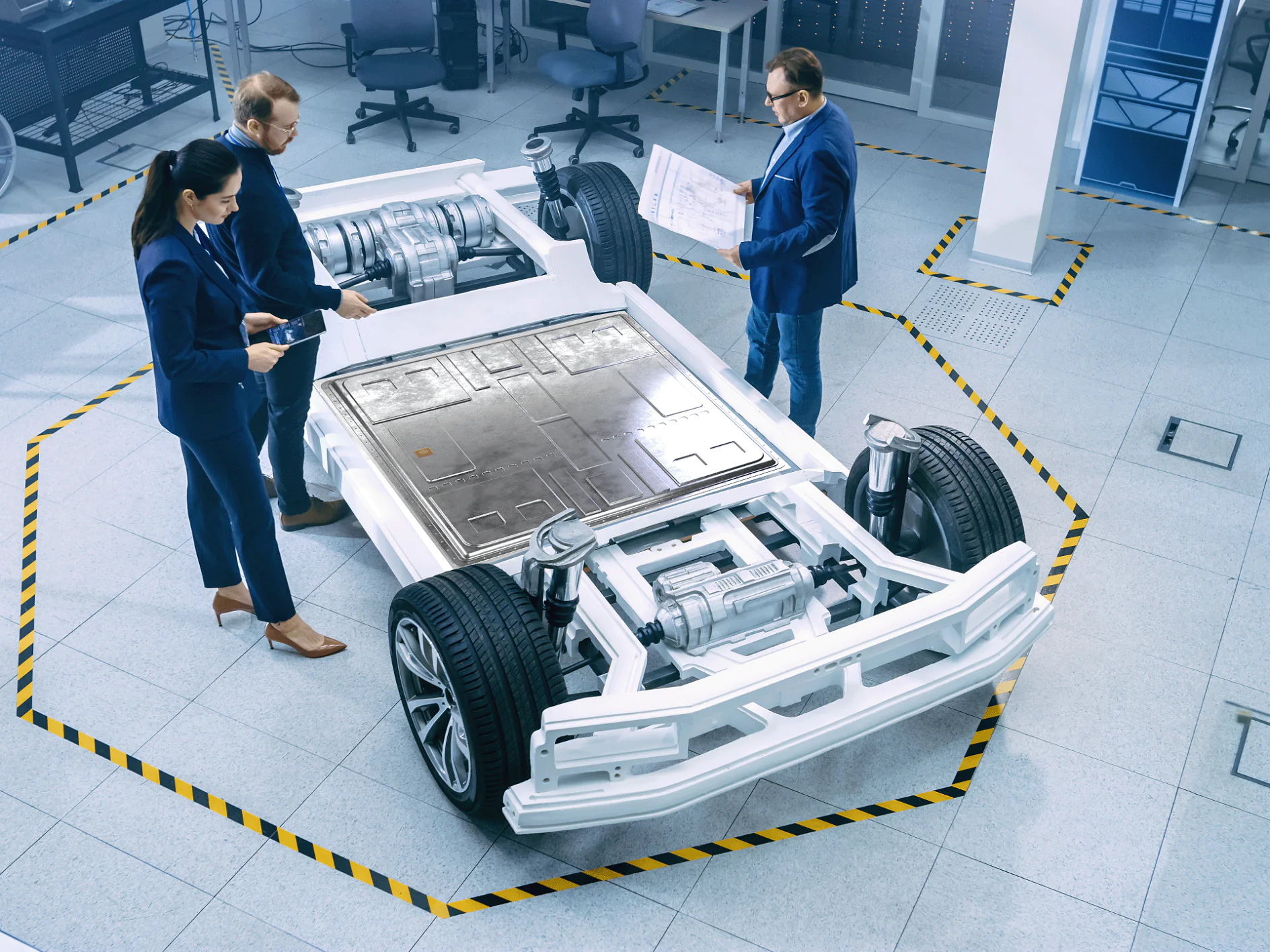 Three people in business attire inspect an electric vehicle chassis in a lab marked with yellow and black tesa tape on the floor. (This text has been generated by AI)