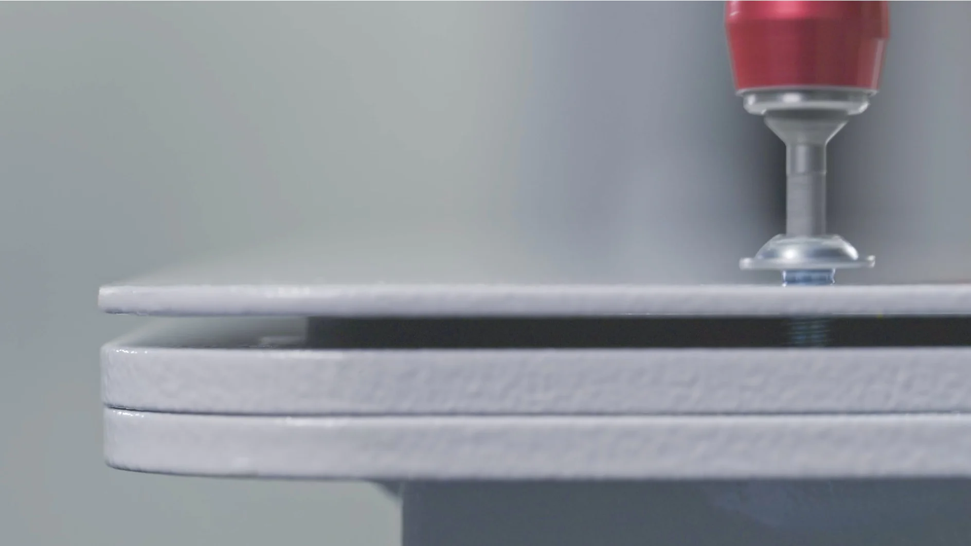 A close-up of a machine tool with a red component attaching two gray metal sheets, held together using tesa tape and a fastener. (This text has been generated by AI)