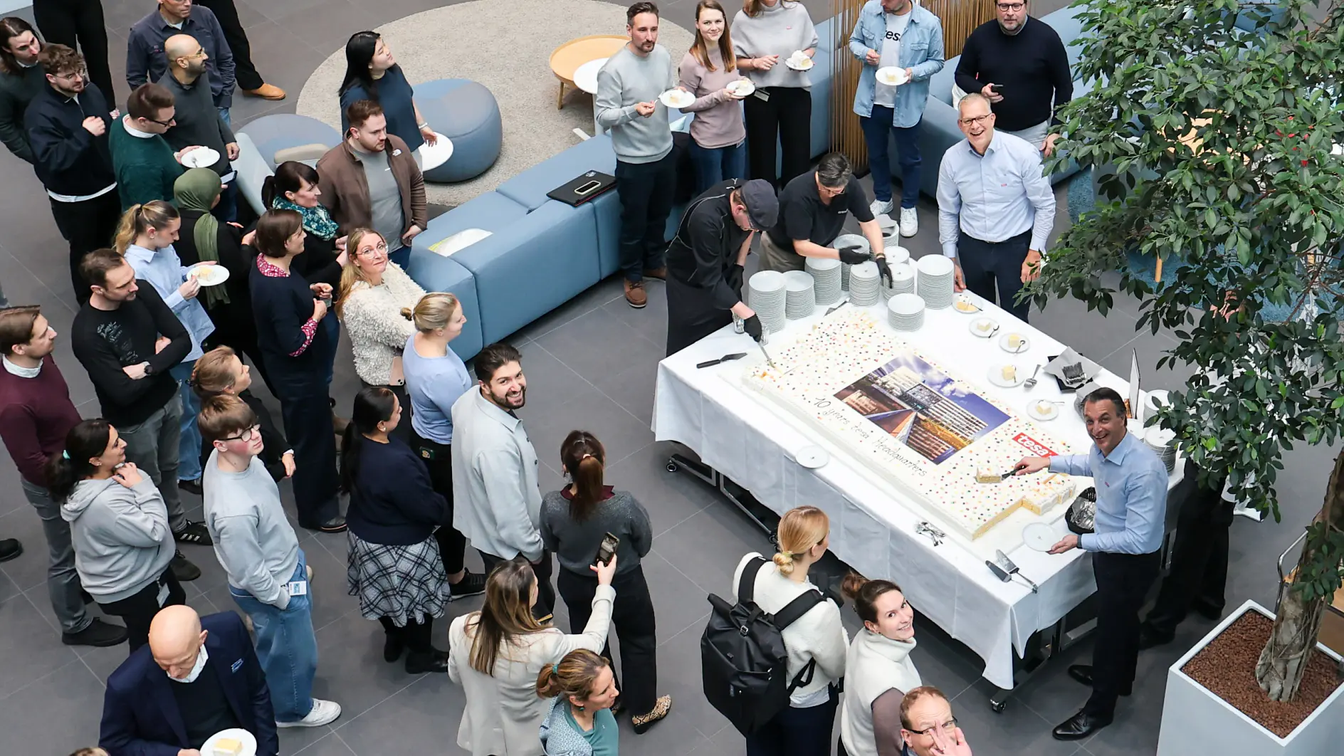 A large group gathers around a table with plates and a big sheet cake in a modern room. No tesa tape is visible in the scene. (This text has been generated by AI)