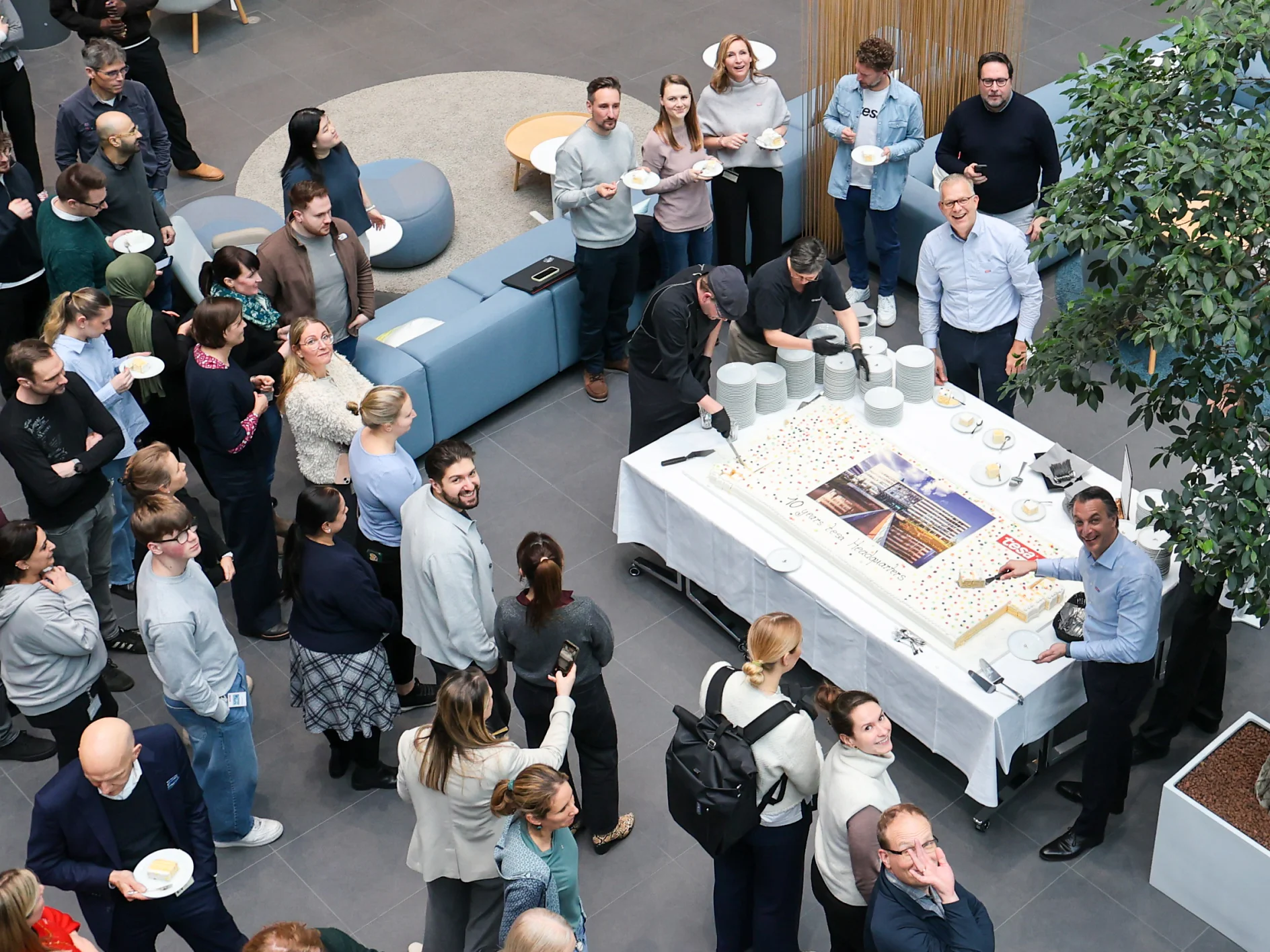 A large group gathers around a table with plates and a big sheet cake in a modern room. No tesa tape is visible in the scene. (This text has been generated by AI)