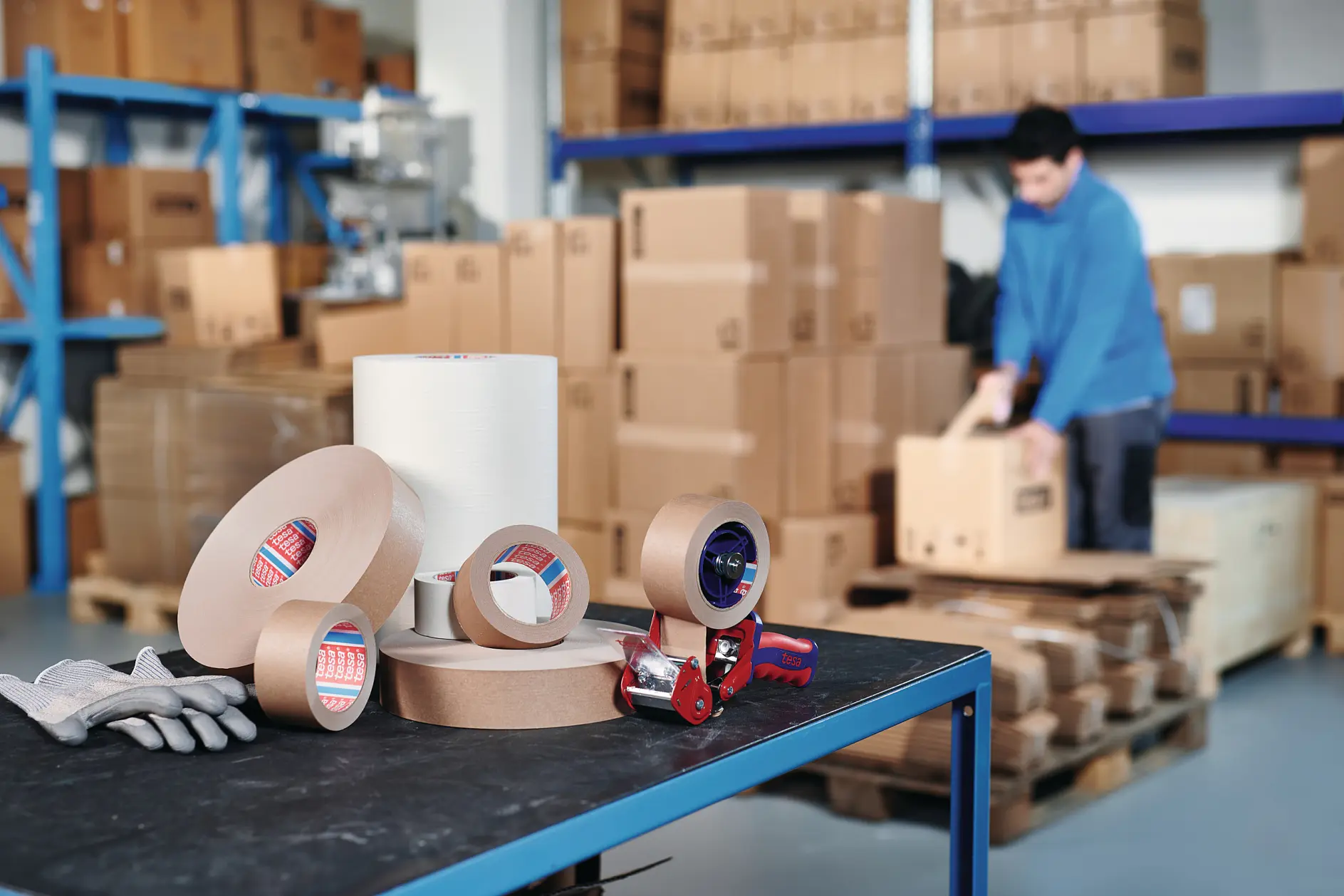warehouse worker stacking cardboard boxes indoors, packing tape rolls and dispenser on table in foreground