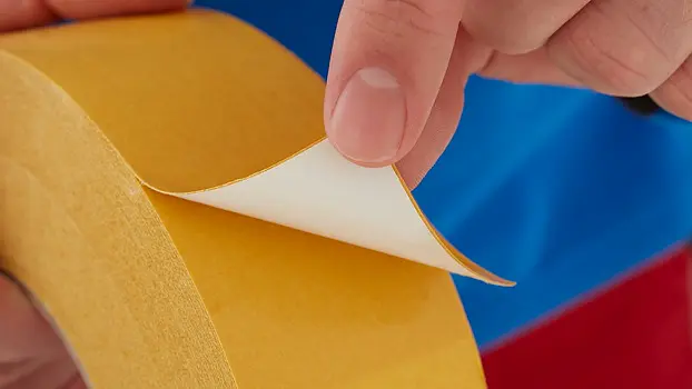 A hand is peeling back the protective paper from a yellow double-sided tesa tape, revealing the adhesive side. The person is wearing a blue and red garment. (This text has been generated by AI)