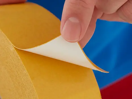 A hand is peeling back the protective paper from a yellow double-sided tesa tape, revealing the adhesive side. The person is wearing a blue and red garment. (This text has been generated by AI)