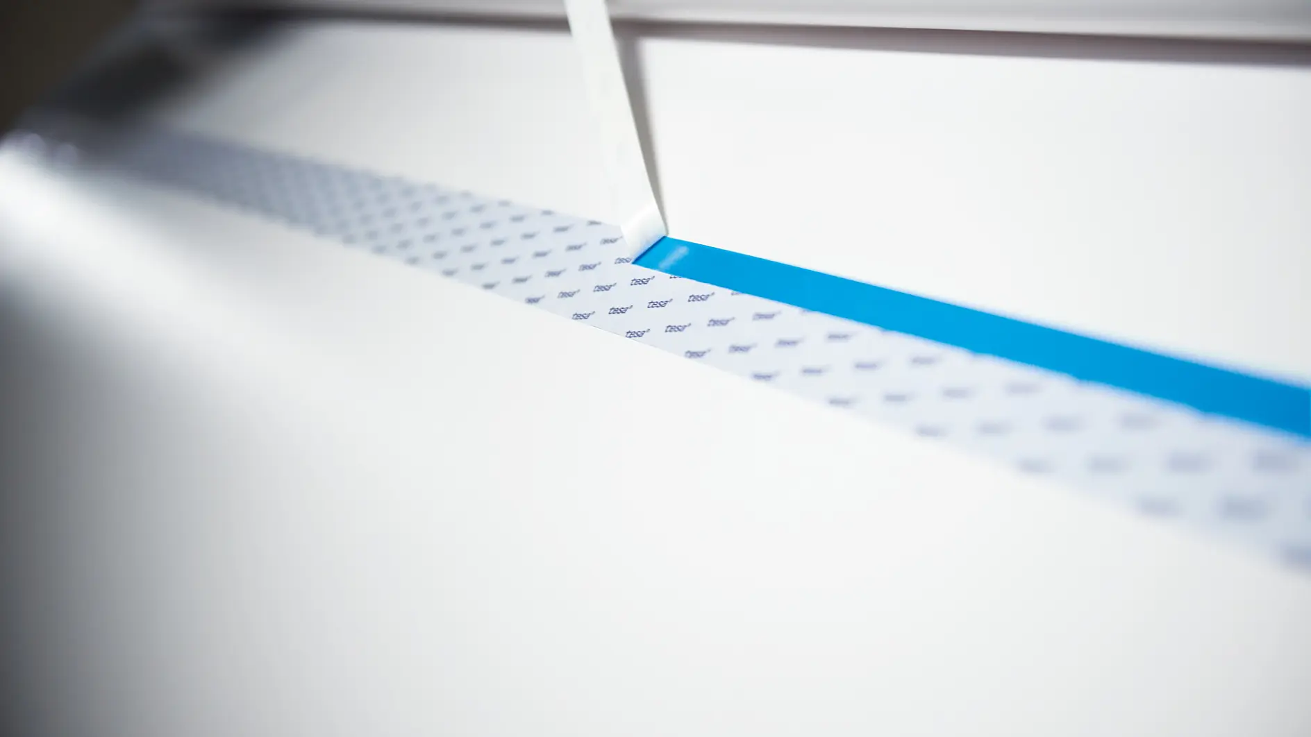 A close-up of a precision cutting tool slicing through paper, revealing a narrow blue strip of tesa tape underneath. The surface being cut appears white with a lightly dotted pattern. The scene is well-lit, emphasizing the sharpness of the cutting edge. (This text has been generated by AI)