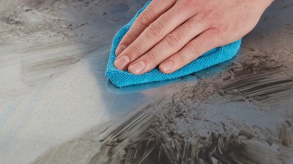 A hand is wiping a dirty surface with a blue microfiber cloth. The surface appears to have smudges and streaks, indicating the cleaning in progress. The focus is on the hand and cloth. (This text has been generated by AI)