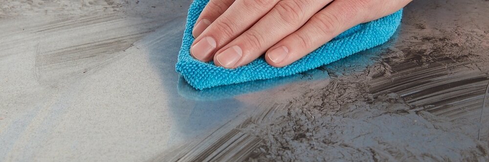 A hand is wiping a dirty surface with a blue microfiber cloth. The surface appears to have smudges and streaks, indicating the cleaning in progress. The focus is on the hand and cloth.