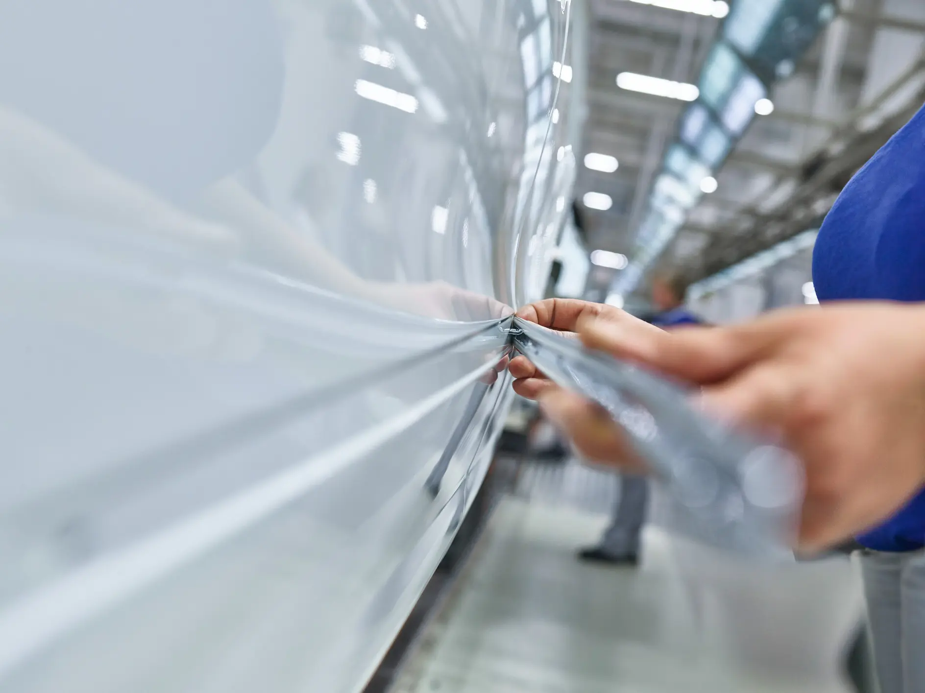 tesa tape being applied to a white vehicle in a factory setting, focusing on hands smoothing the tape along the surface (This text has been generated by AI)