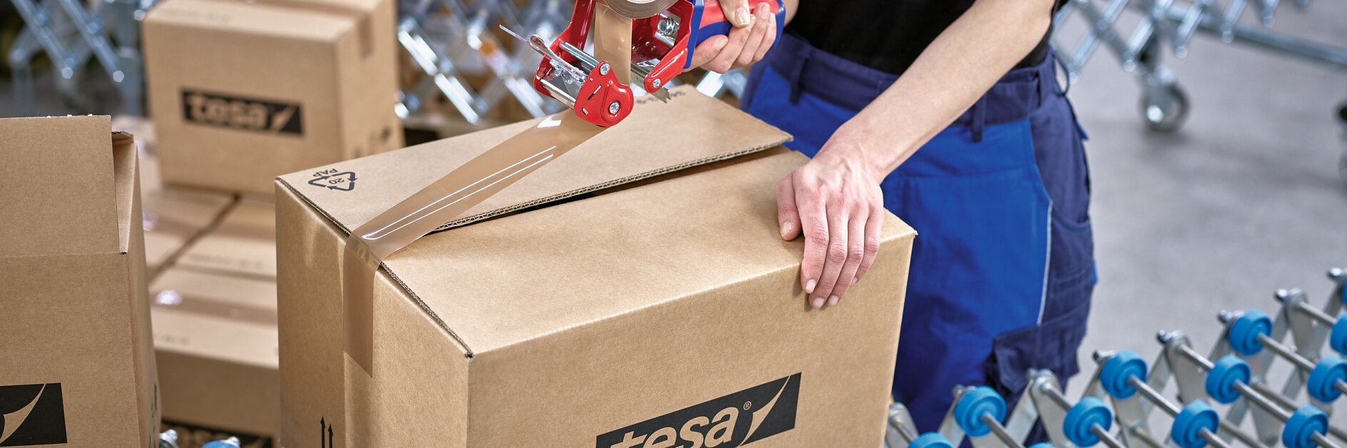 person sealing a tesa-branded cardboard box with tape dispenser on a conveyor belt in a warehouse setting