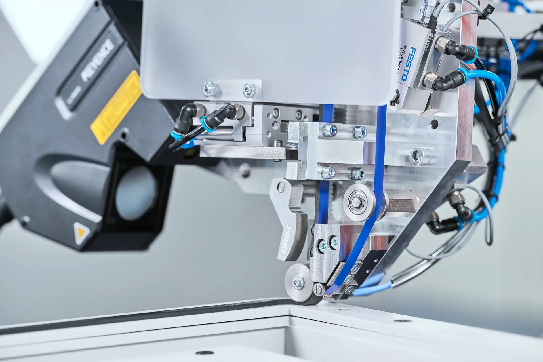 A close-up of an industrial machine with metal parts, blue cables, a sensor, and tesa tape near a white surface in a factory setting. (This text has been generated by AI)