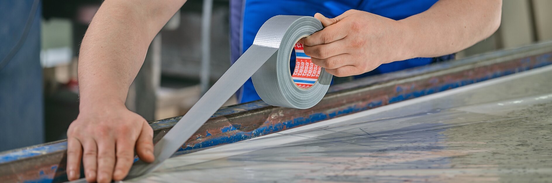 tesa worker in gray shirt and blue pants applying silver duct tape to a large flat surface in an indoor workshop setting