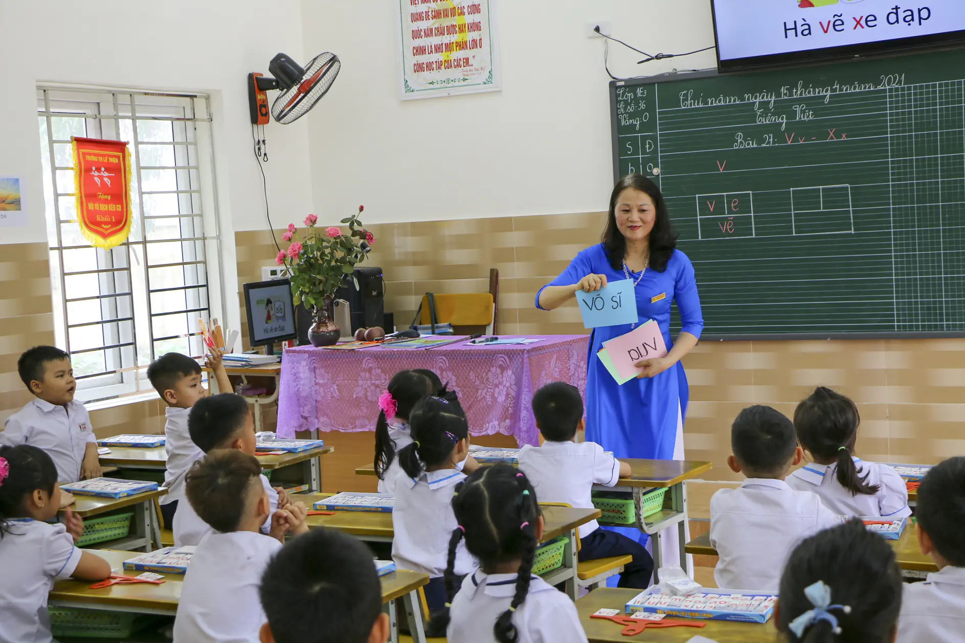 A teacher in a blue dress uses tesa tape to hold up vocabulary cards, as students in white uniforms watch from their desks. (This text has been generated by AI)