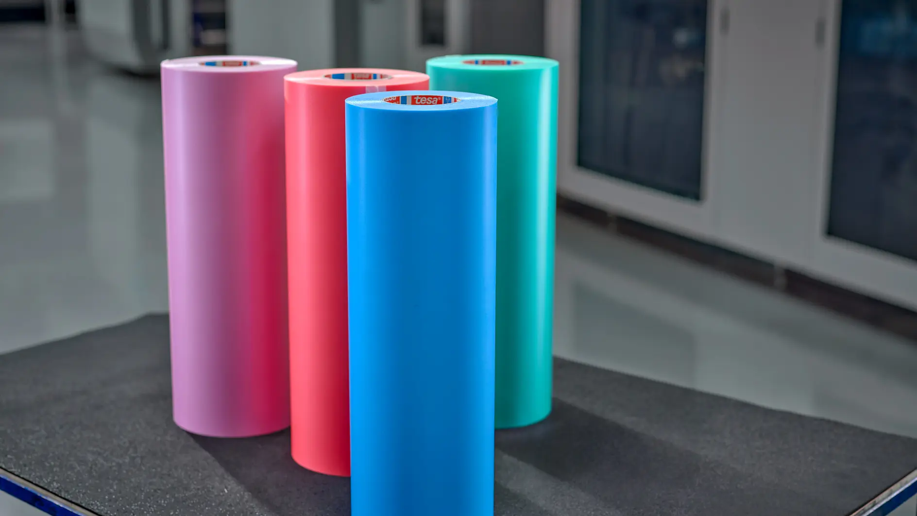 Five rolls of colorful tesa adhesive tape in pink, red, blue, green, and gray, displayed on a black surface indoors. (This text has been generated by AI)